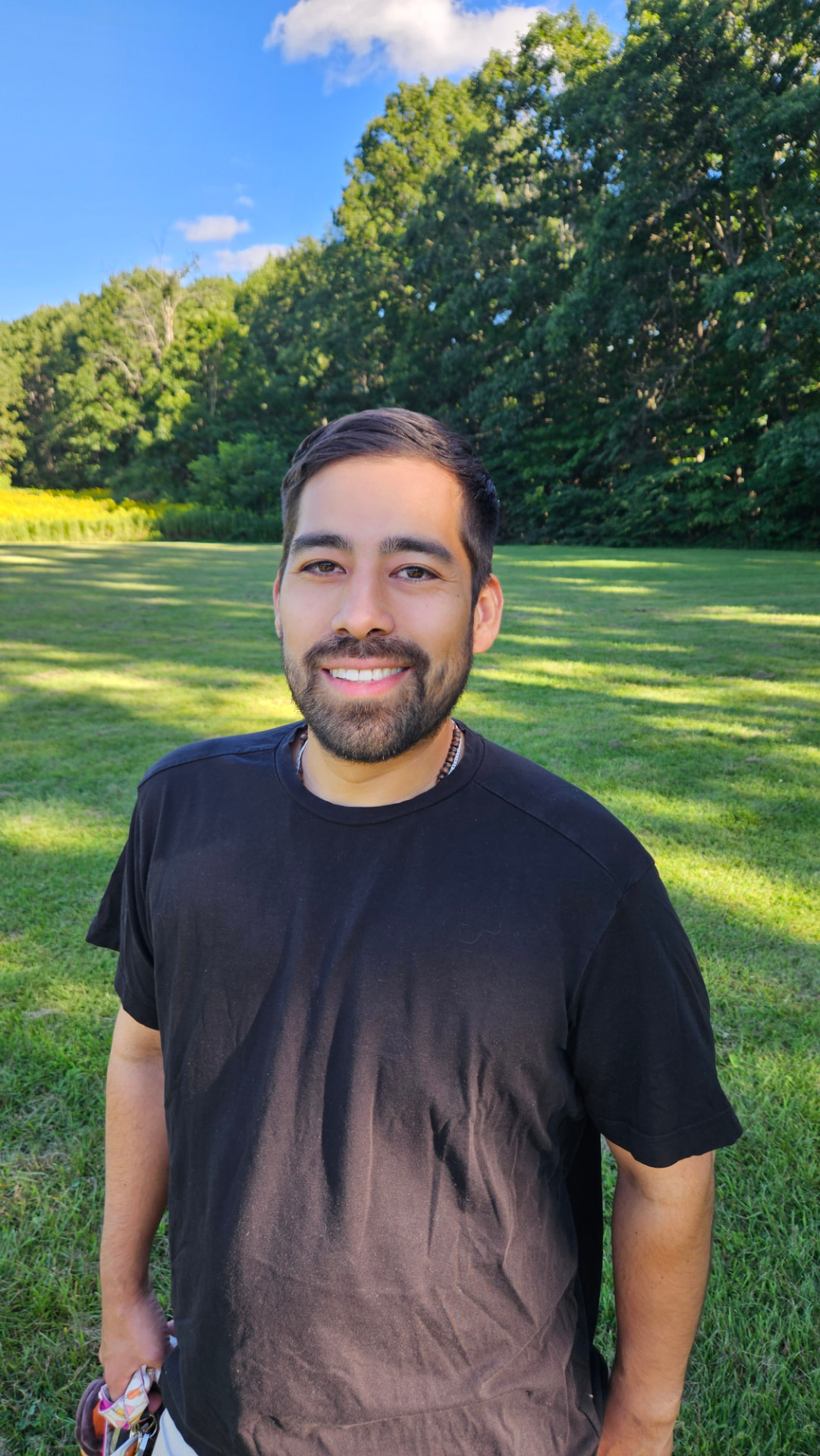 Nolan Fernandez smiling outdoors in a grassy meadow with trees behind him.